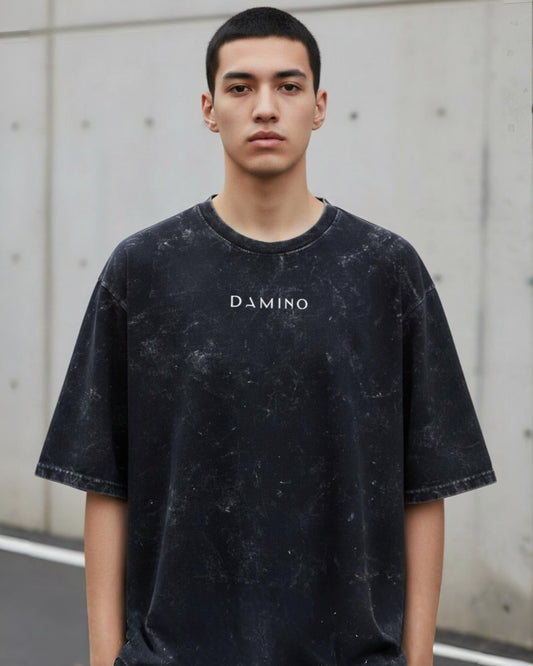 Person wearing a black t-shirt with 'DAMINO' logo against a concrete wall.